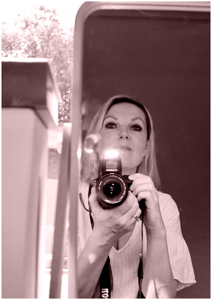 Pale pink tint on A2 portrait sized black and white image of a woman reflected in a vanity mirror sat holding a black canon camera with flash operated. The woman wears a white and pink striped v-neck blouse. To the left of the mirror, the edge of a box is visible and in the distance there is a tree.