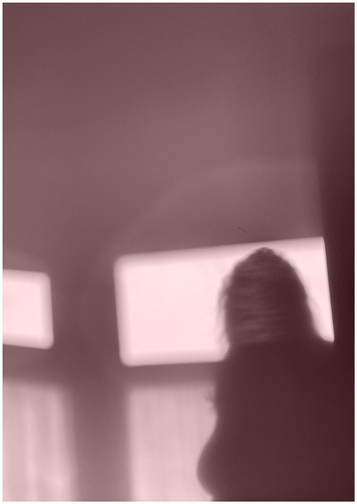 Pale pink tint on A2 portrait sized black and white image of the silhouette of the top half of a woman’s body stood in front of a window with daylight showing through four window panes.
