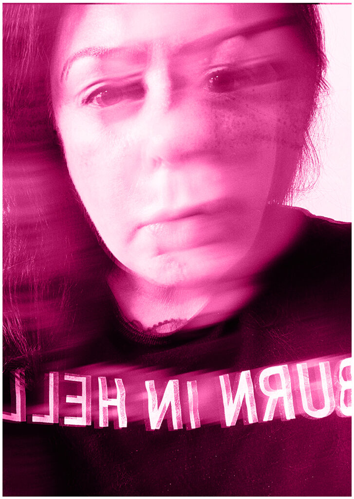 Hot pink tint on A2 portrait sized black and white image of blurred head and shoulders photograph of a woman’s face facing the camera. She wears a dark top with the words ‘BURN IN HELL’ printed in white across the chest. The words are shown in reverse as if reading in a mirror.