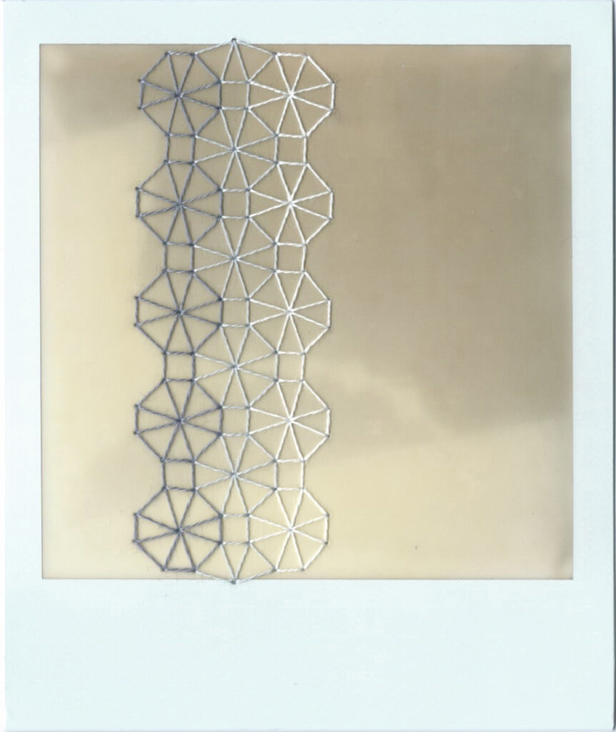 An undeveloped polaroid that has been stitched down the left hand side using a traditional blackwork embroidery pattern. The stitching is done in gradients of grey. The pattern consists of three columns of an eight sided, segmented shape that looks a bit like a wheel with spokes.